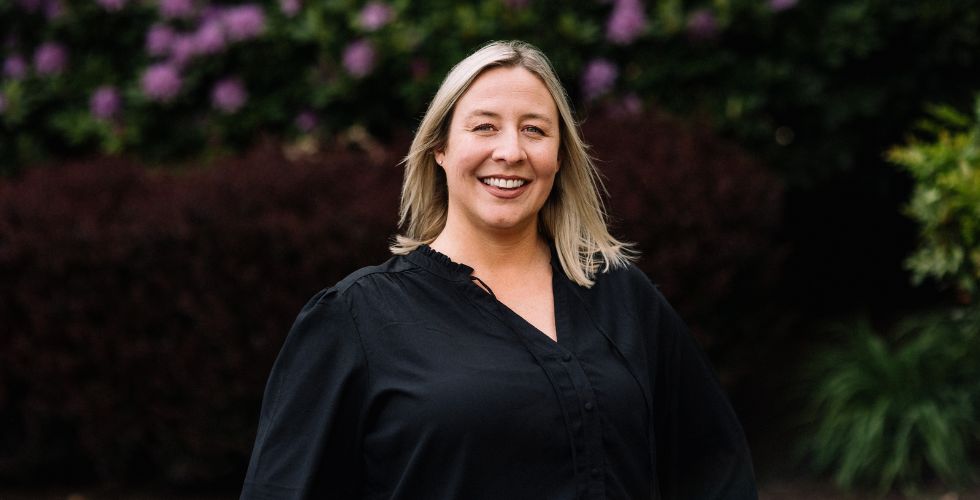 A woman with blonde hair wearing a black blouse smiles while standing outdoors in front of bushes and greenery.