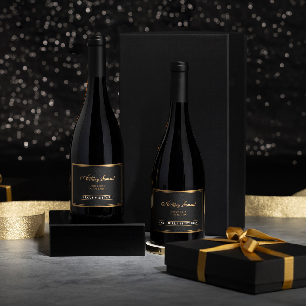 Two elegant Pinnacle Pinot Noir wine bottles with black and gold labels are displayed before a black gift box, alongside a small black present tied with a gold ribbon. A gold ribbon and sparkly background add a festive touch.