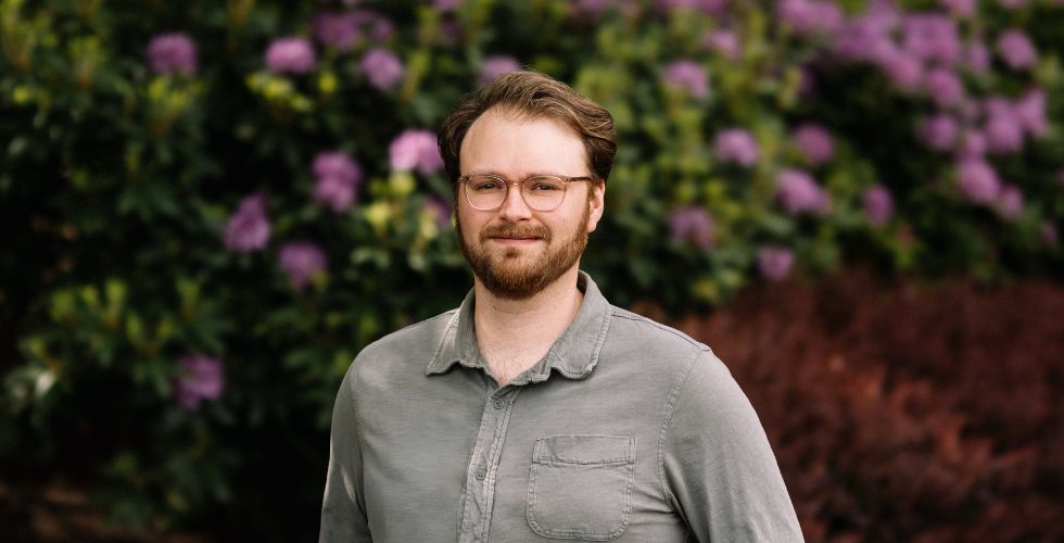 A man with glasses and a beard stands outdoors, smiling, wearing a light gray button-up shirt. The background features green foliage and blooming purple flowers.