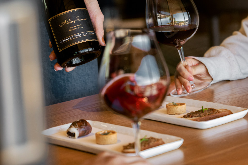 A person pours red wine from a bottle into a glass at a table set with small gourmet appetizers on white plates. Another hand holds up a filled wine glass.