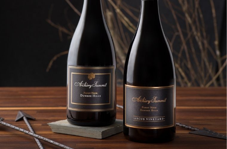 Two bottles of Archery Summit Pinot Noir wine are displayed on a dark surface, with decorative arrows and bare branches in the background, creating a rustic and elegant setting.