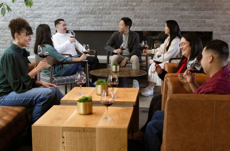 Seven people sit in a cozy lounge, chatting and smiling while holding glasses of red wine. They are gathered around wooden coffee tables with small potted plants, in a relaxed, modern setting.