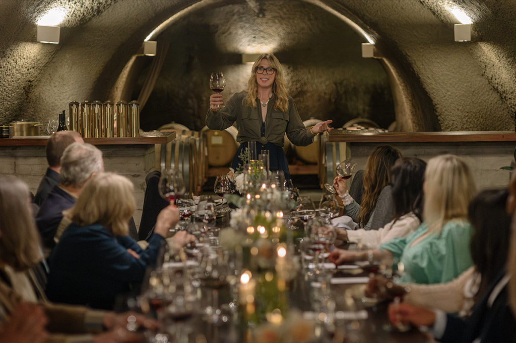 A woman stands at the head of a long candlelit table in a wine cellar, leading an Epicurean Experience: Harvest Dinner as she raises her glass and invites guests on either side to join her in a toast.