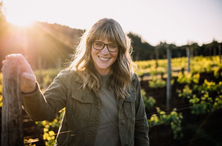 A woman with long blonde hair and glasses, wearing a green jacket, smiles while standing in a sunlit vineyard during golden hour. Rows of grapevines and posts are visible in the background.