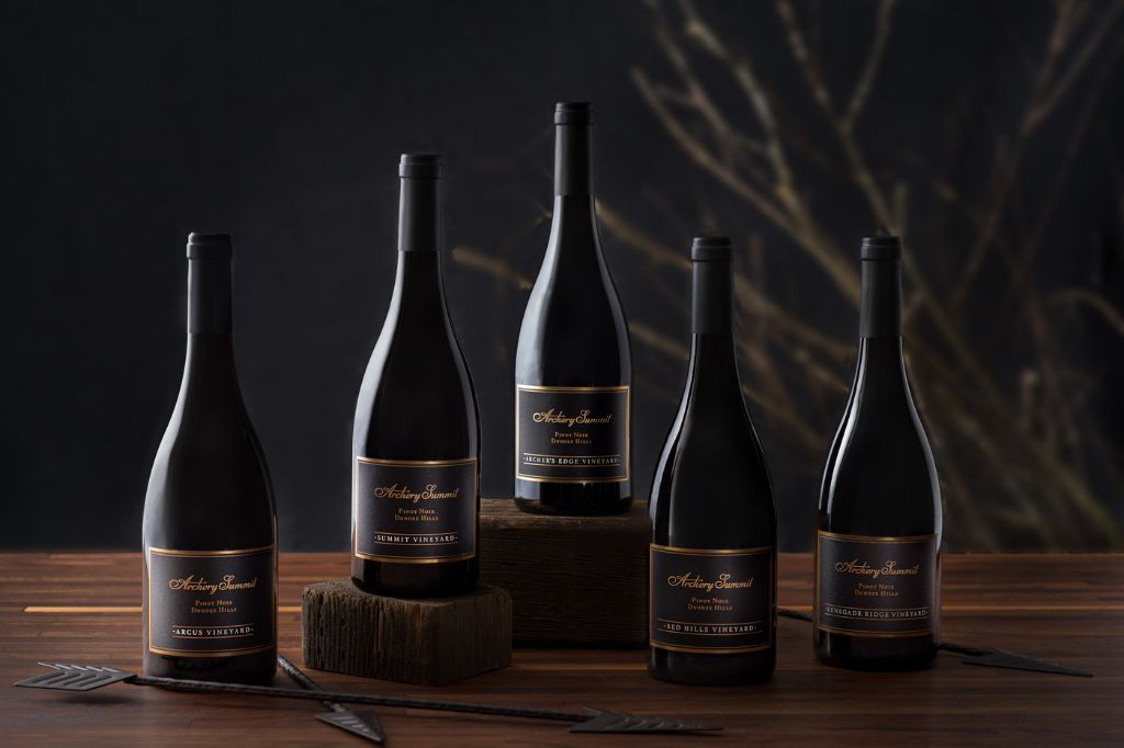 Five dark wine bottles with elegant black and gold labels are arranged on a wooden surface, some elevated on wood blocks, with decorative arrows and blurred branches in the background.