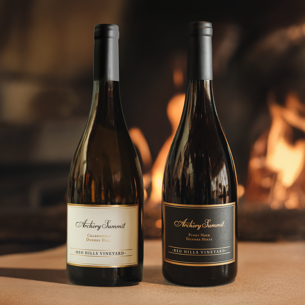 Two bottles of Archery Summit wine, including the Pinnacle Pinot Noir Wine Gift and a Chardonnay, are placed side by side on a table, with a warm, glowing fireplace softly blurred in the background.