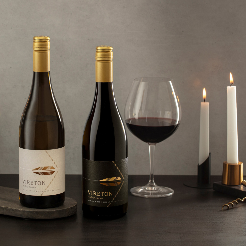 Two bottles of Vireton wine from the 93 Point Stunners 3-Bottle Collection sit on a dark table beside a large glass of red wine, two lit candles, and a corkscrew, with a neutral gray background creating a sophisticated ambiance.