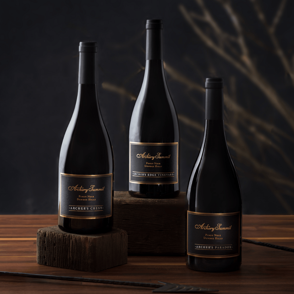 Three bottles from the 93 Point Stunners 3-Bottle Collection of Archery Summit Pinot Noir are displayed on wooden blocks against a dark, blurred background with subtle branch patterns. Each bottle features a black label with gold accents.