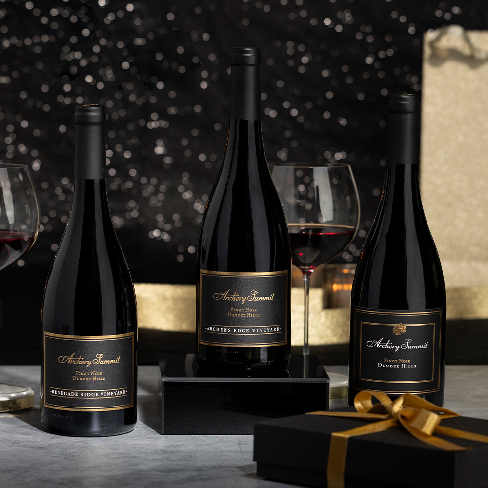 Three bottles of Archery Summit Pinot Noir—part of the 93 Point Stunners 3-Bottle Collection—are displayed with two glasses of red wine, a gold gift box with a black ribbon, and a glittery black background.