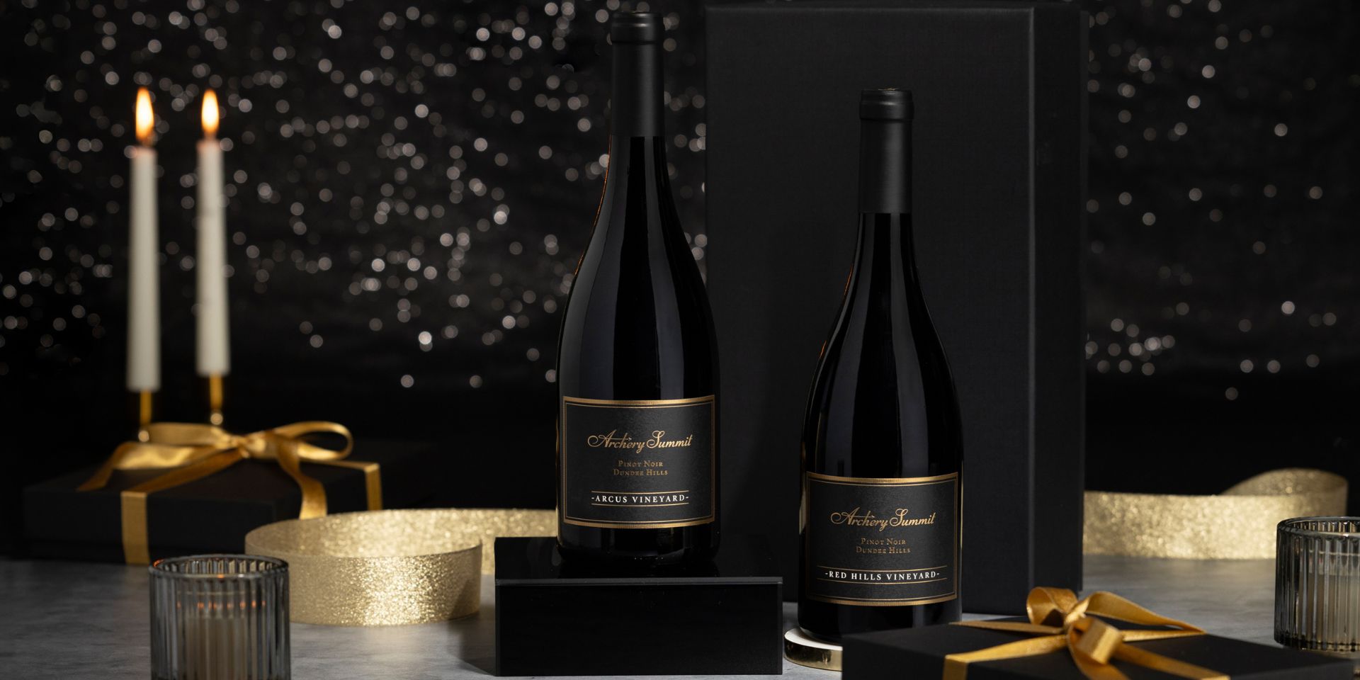 Two elegant wine bottles with black and gold labels are displayed with black gift boxes, gold ribbon, lit candles, and a candle holder, set against a dark, sparkling background.