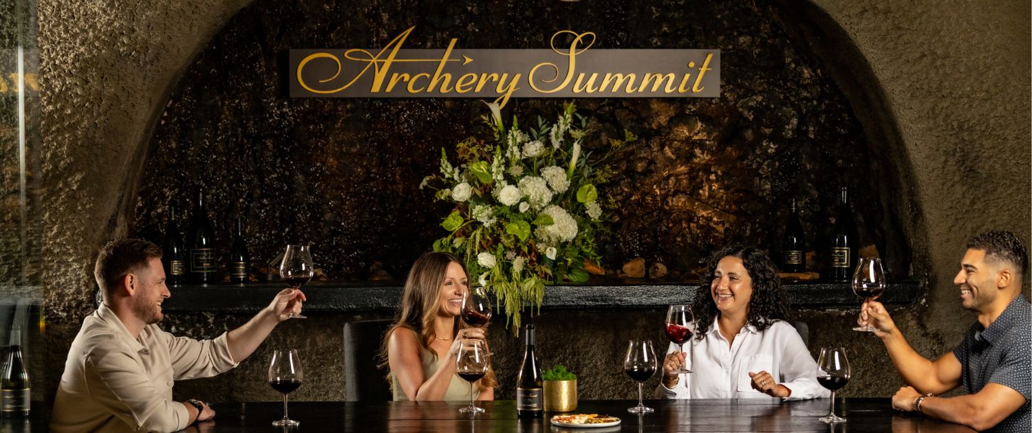 Four people sit around a table in a dimly lit wine cellar, raising glasses of red wine in a toast. Behind them is a large floral arrangement and a sign reading Archery Summit.