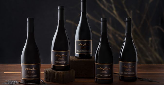 Five dark wine bottles with black and gold labels are arranged on a wooden surface, some elevated on blocks, against a dark, blurred background with subtle decorative twigs—perfect for those looking to buy wine with elegance in mind.