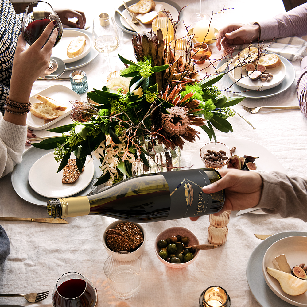 A table set for a meal with plates of bread, crackers, olives, and dips. A person pours Vireton Pinot Gris while others hold glasses. A floral centerpiece decorates the table.