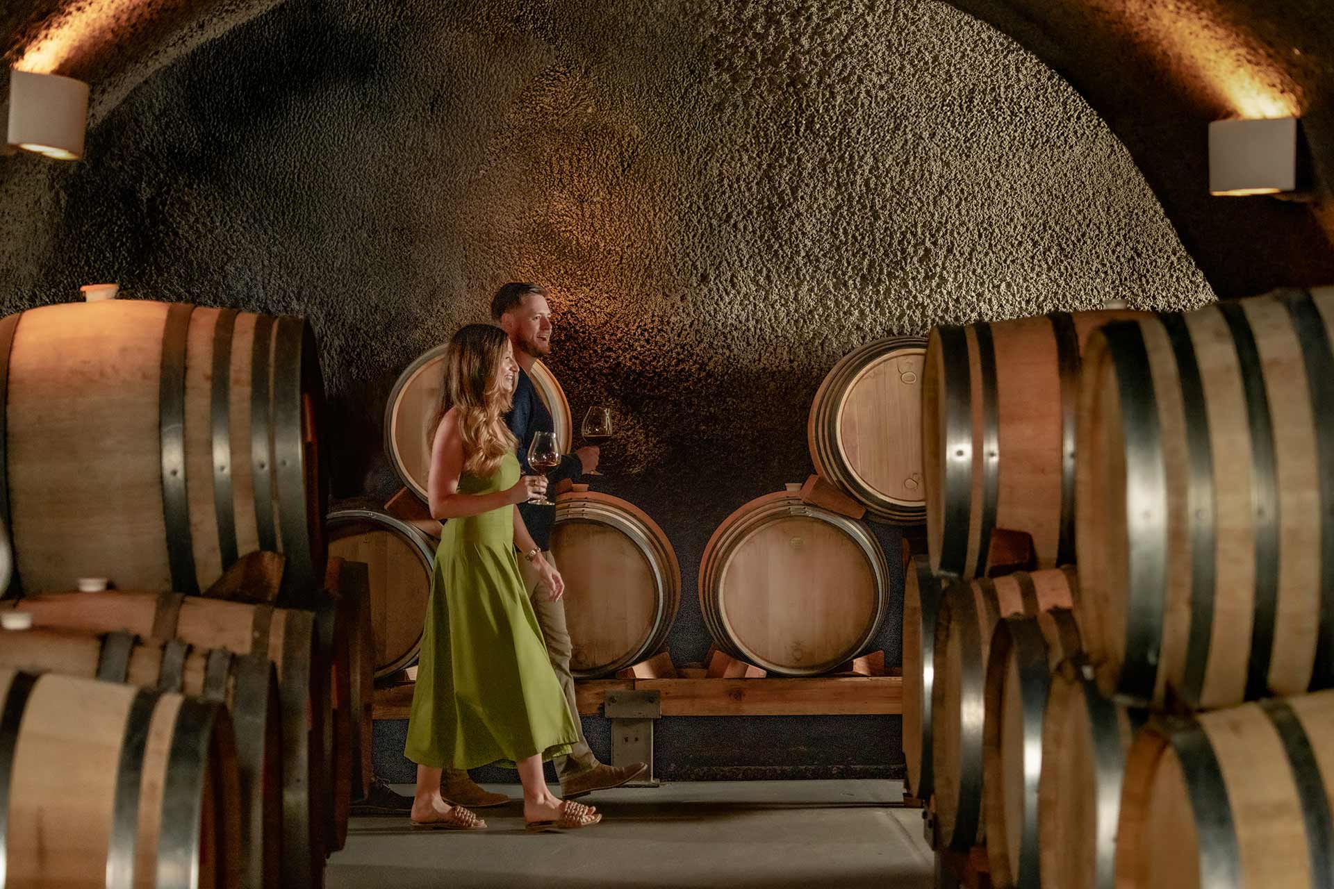 A man and woman holding wine glasses walk through a wine cellar lined with large wooden barrels, under arched ceilings with textured walls.
