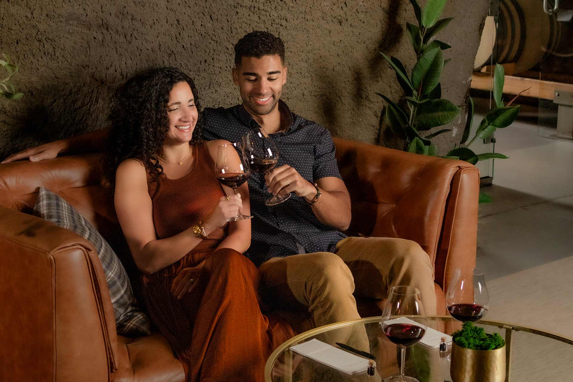 A smiling couple sits on a brown leather couch, clinking wine glasses. They are relaxed and enjoying each others company, with plants and a glass table with more wine glasses nearby.