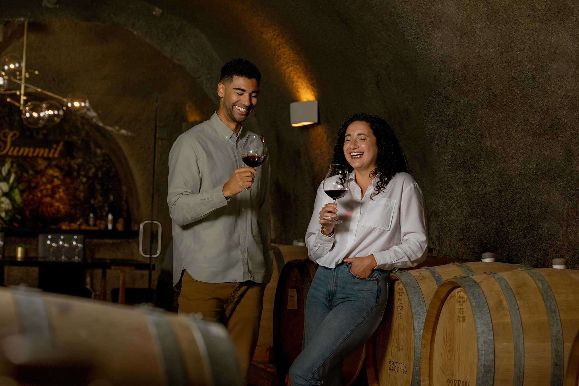 A man and woman stand in a wine cellar, smiling and holding glasses of red wine. They are leaning against wooden wine barrels and appear to be enjoying a pleasant conversation.