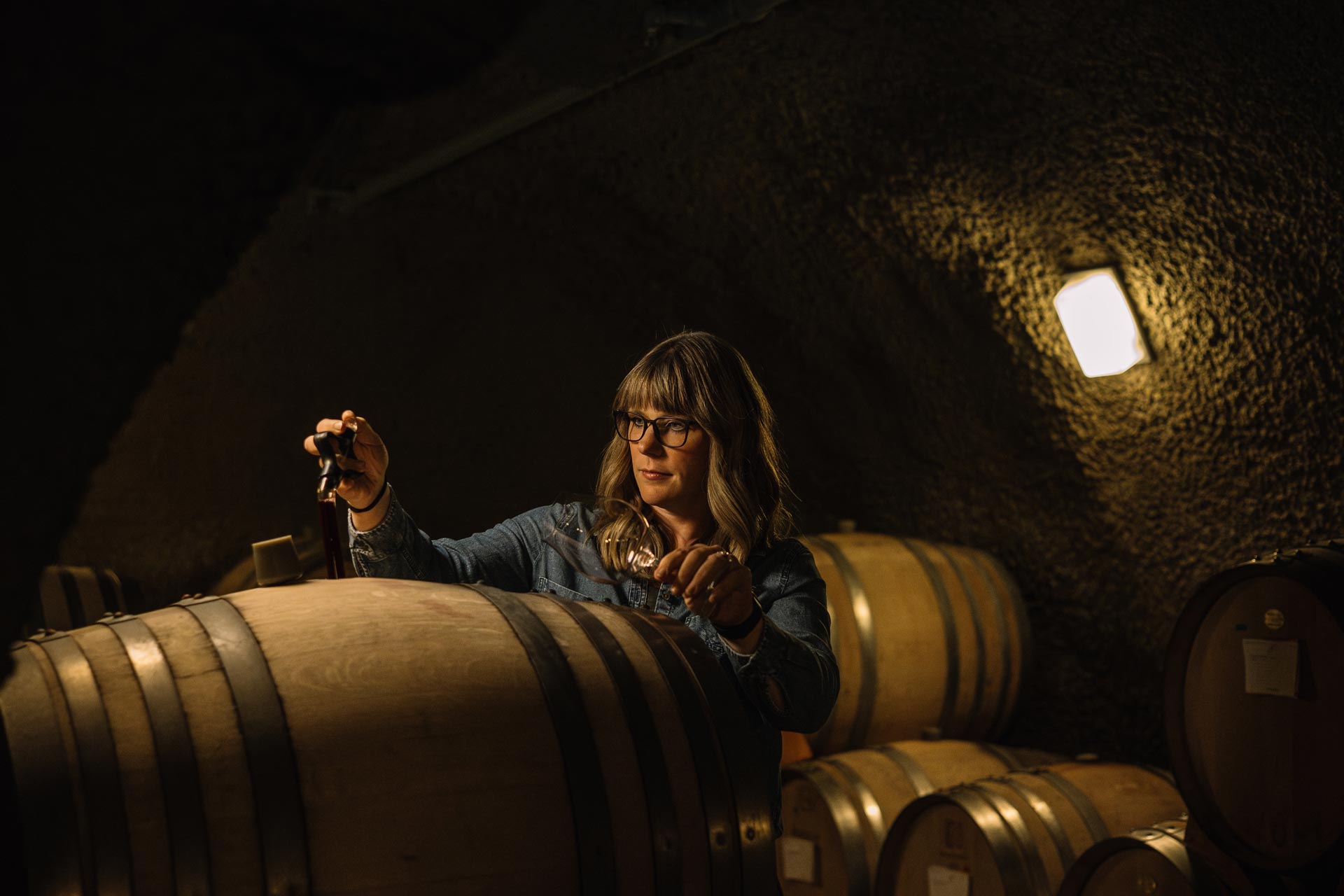 A woman with long hair and glasses takes a wine sample from a wooden barrel in a dimly lit wine cellar, surrounded by more barrels stacked in the background.