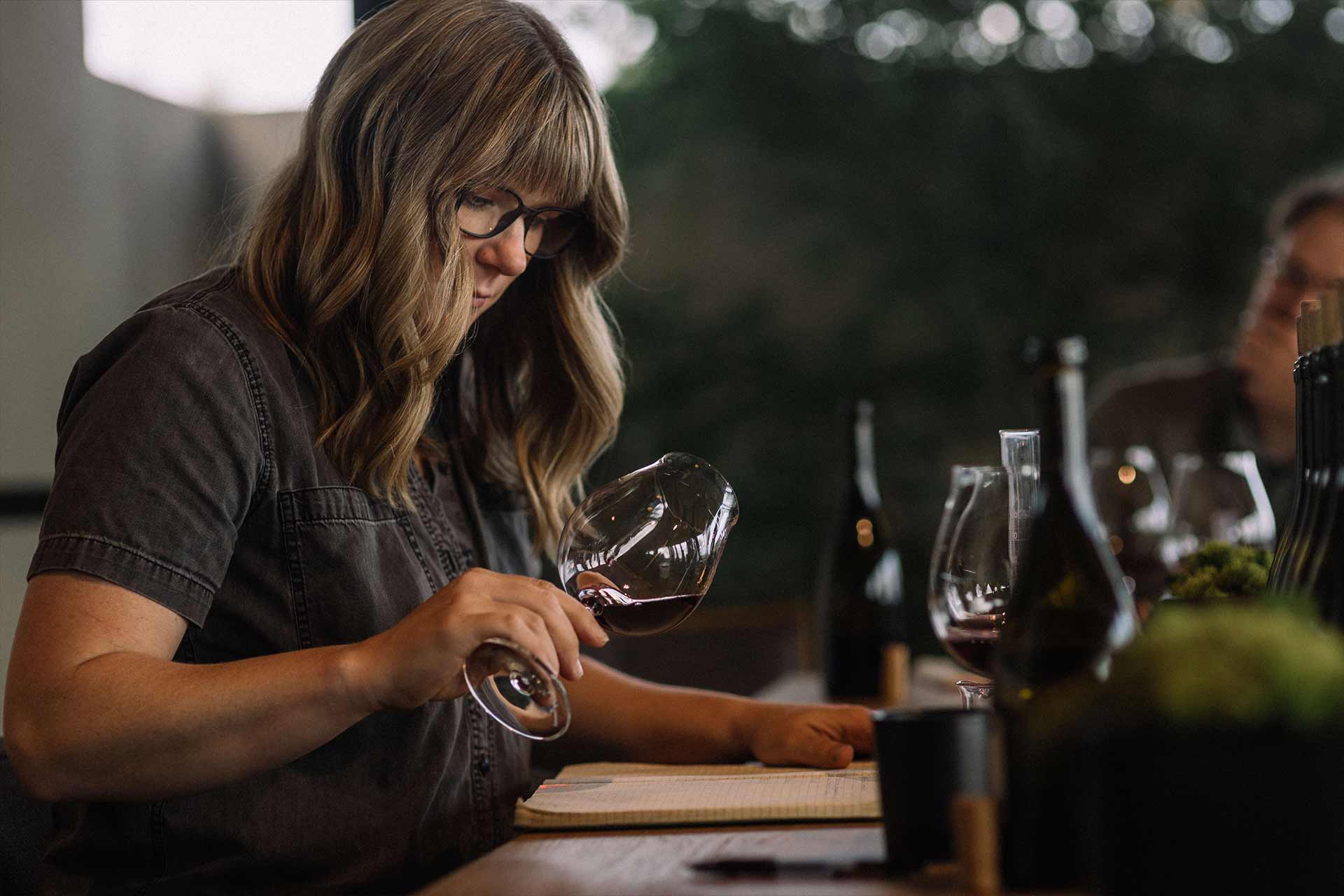 A person with long hair and glasses examines a glass of red wine while sitting at a table with wine bottles and glasses, appearing focused and engaged in tasting or evaluation.