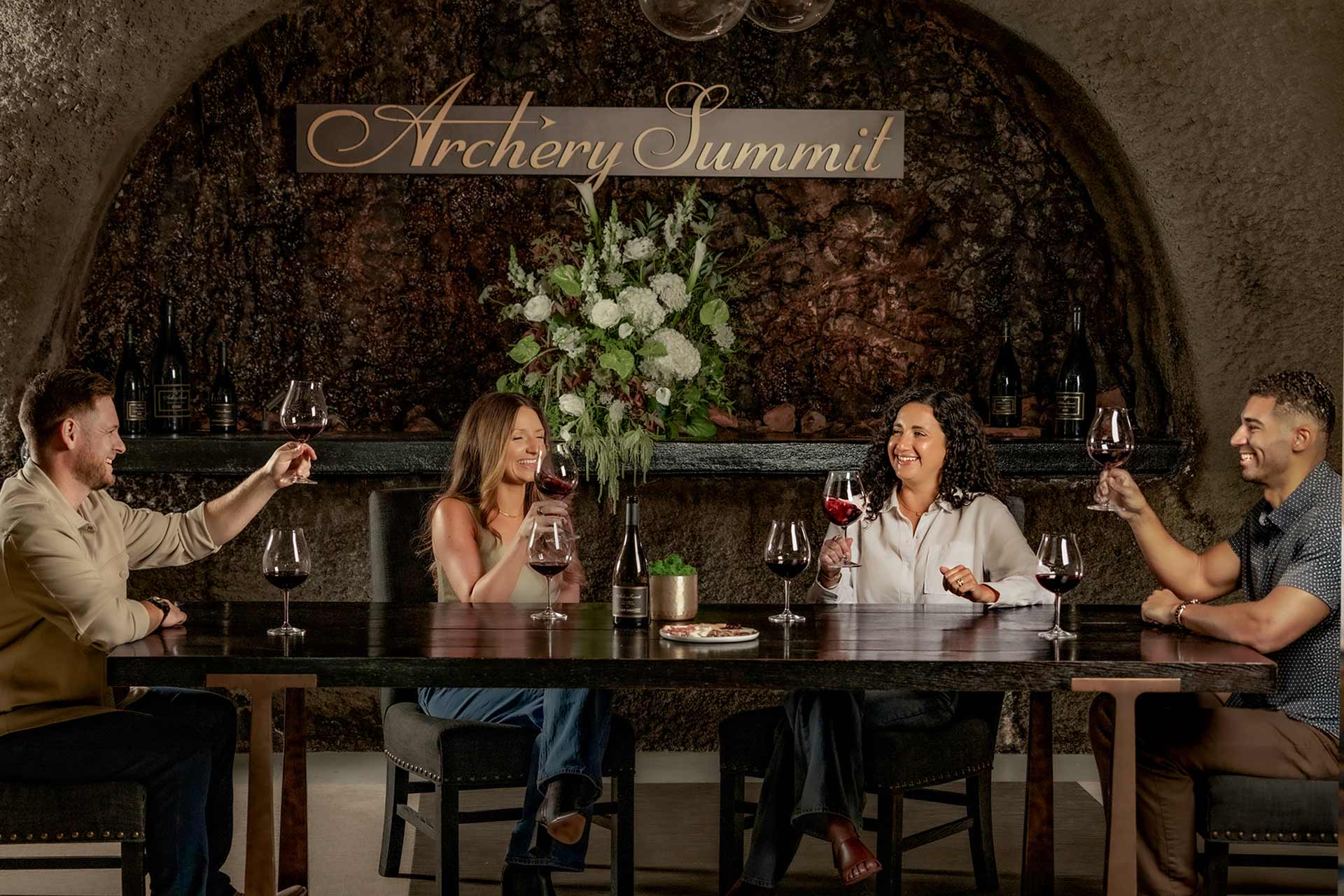 Four people sit around a table, smiling and raising glasses of red wine in a toast. Behind them is a floral arrangement and a sign that reads Archery Summit. The setting looks cozy and elegant.