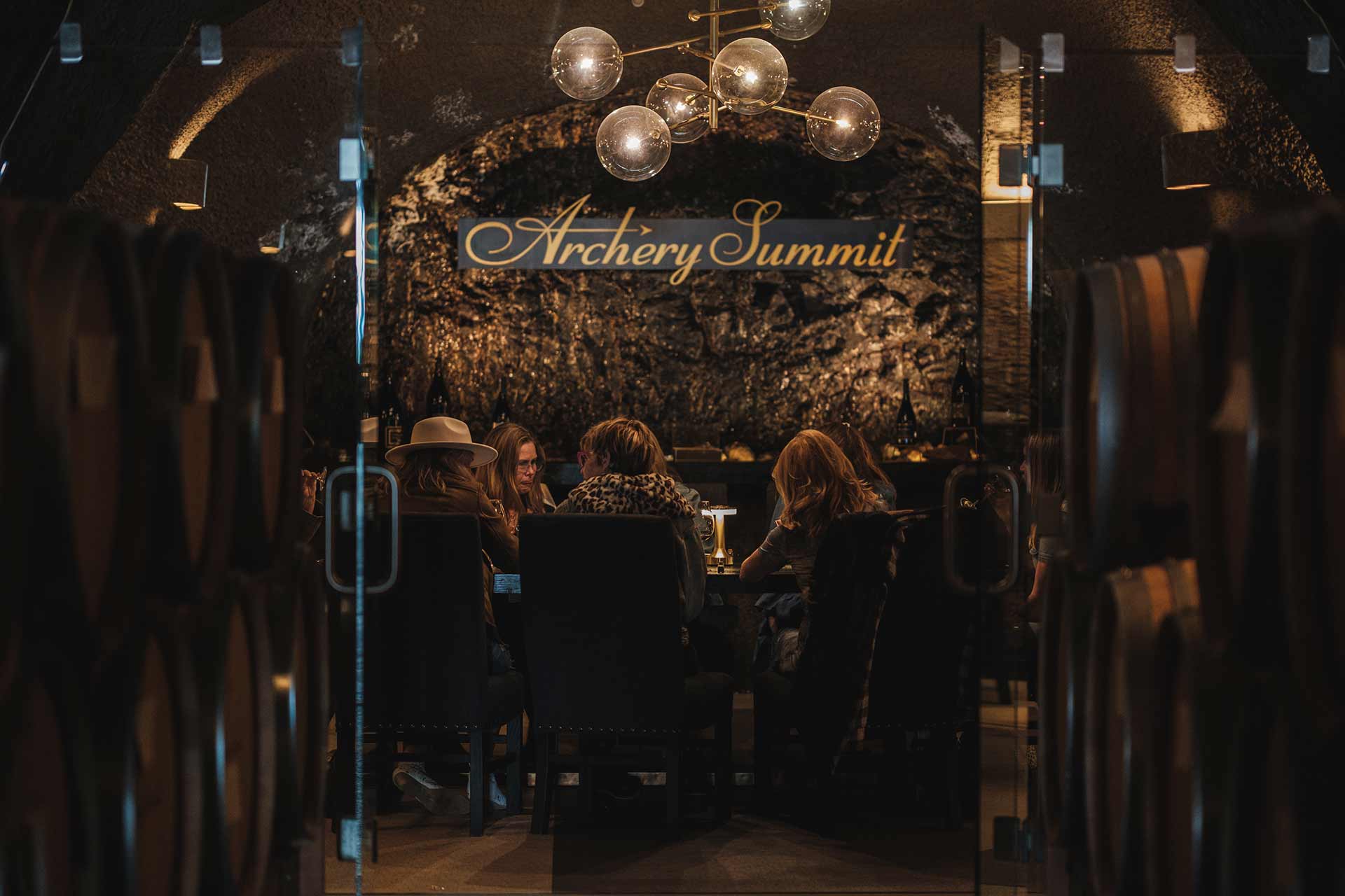 A group of people sit around a table inside a dimly lit wine cellar at Archery Summit, surrounded by wine barrels and illuminated by a modern chandelier.
