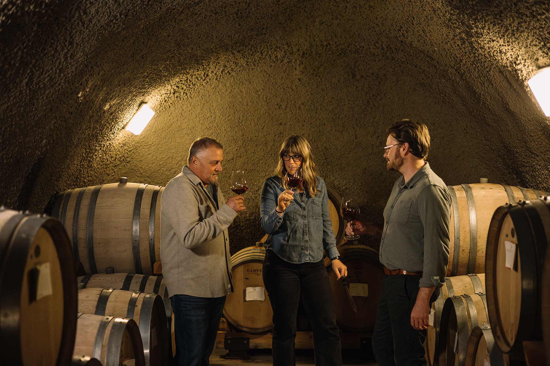 Three people stand in a dimly lit wine cellar, surrounded by wooden barrels, holding wine glasses and tasting wine together.