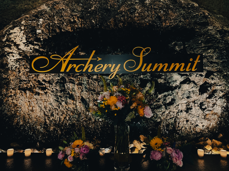 A rocky backdrop with Archery Summit in elegant yellow script, a floral arrangement in a vase, smaller bouquets, and lit candles evoke the Epicurean Experience: Alchemy of Food in an enchanting display.