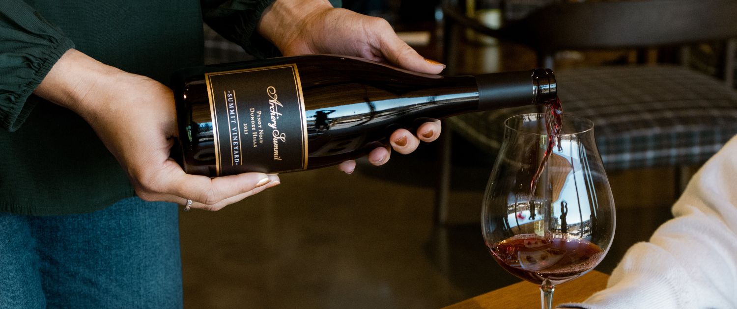 A person pours red wine from a bottle labeled Anthony Summit into a large wine glass on a wooden table, with a blurred indoor setting in the background.