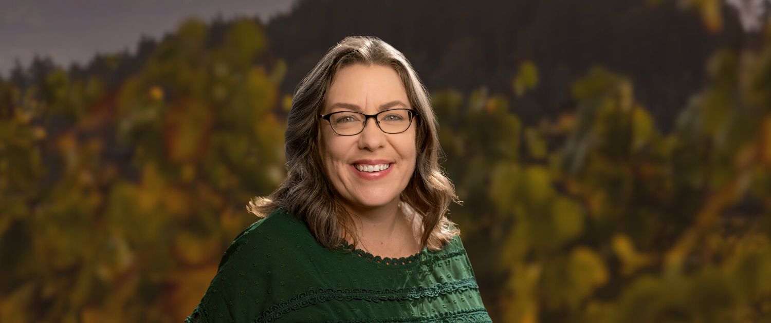 A woman with wavy brown hair and glasses smiles at the camera. She is wearing a green blouse and standing outdoors in front of a blurred background of green trees and foliage.