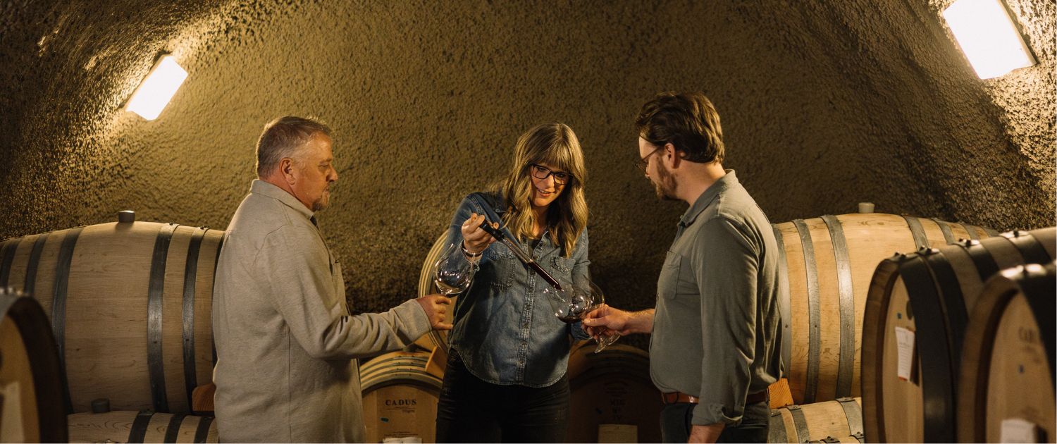 Three people stand in a wine cellar surrounded by barrels; a woman in the center is pouring wine into a glass held by a man, while another man watches. Soft lighting creates a warm, intimate atmosphere.