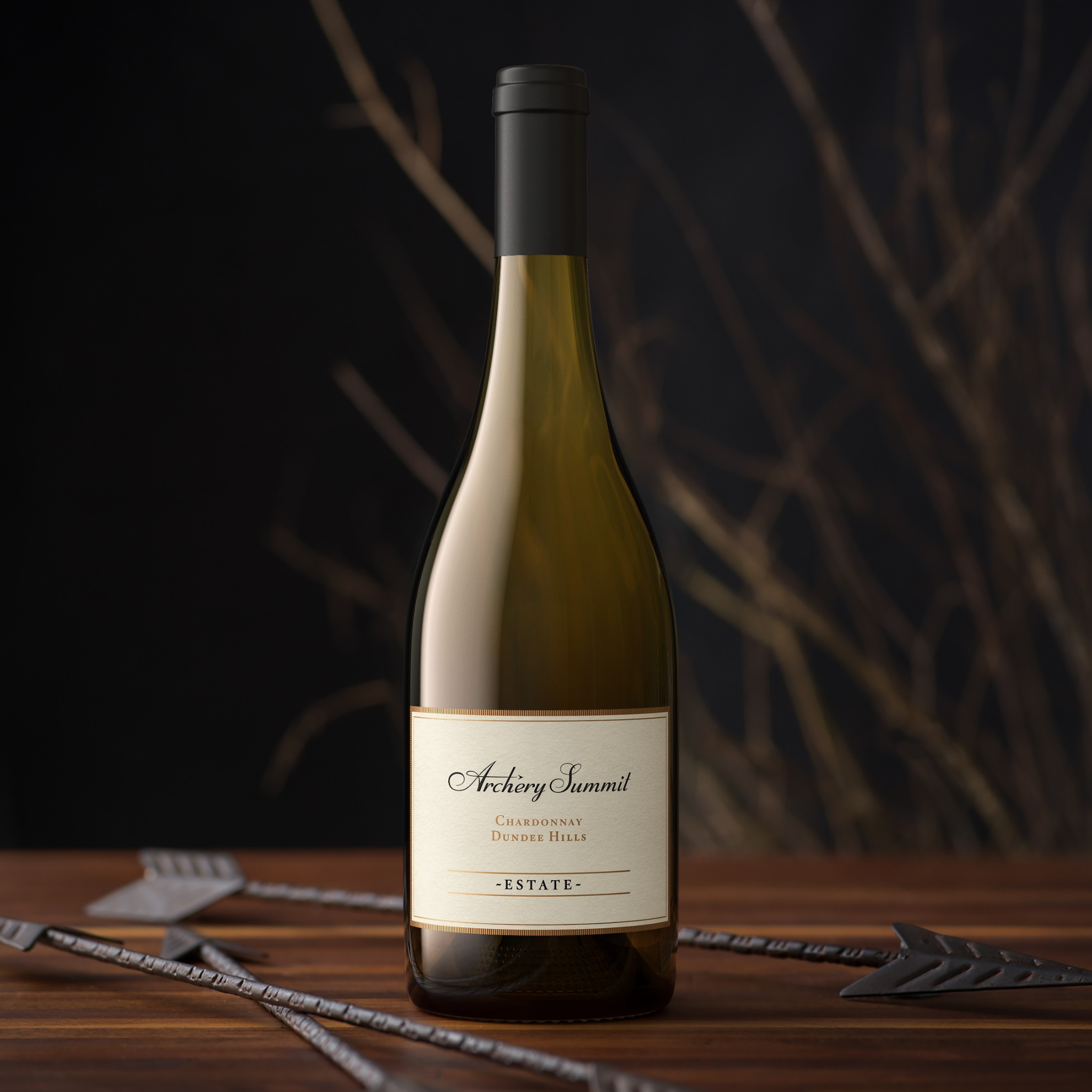 A bottle of Dundee Hills Estate Chardonnay by Archery Summit stands on a wooden surface with metal arrows beside it. The dark background features blurred bare branches, creating an elegant and atmospheric scene.
