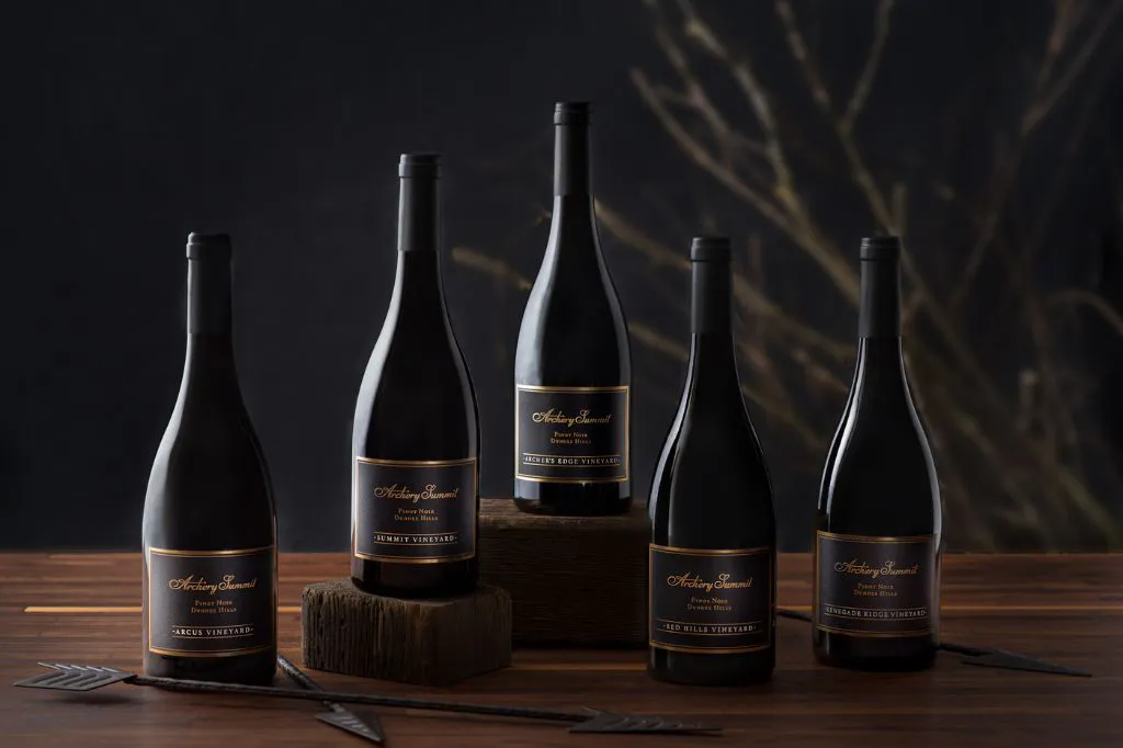 Five dark wine bottles with black and gold labels are displayed on a wooden surface, with some bottles elevated on wooden blocks. The background is dark with out-of-focus tree branches.