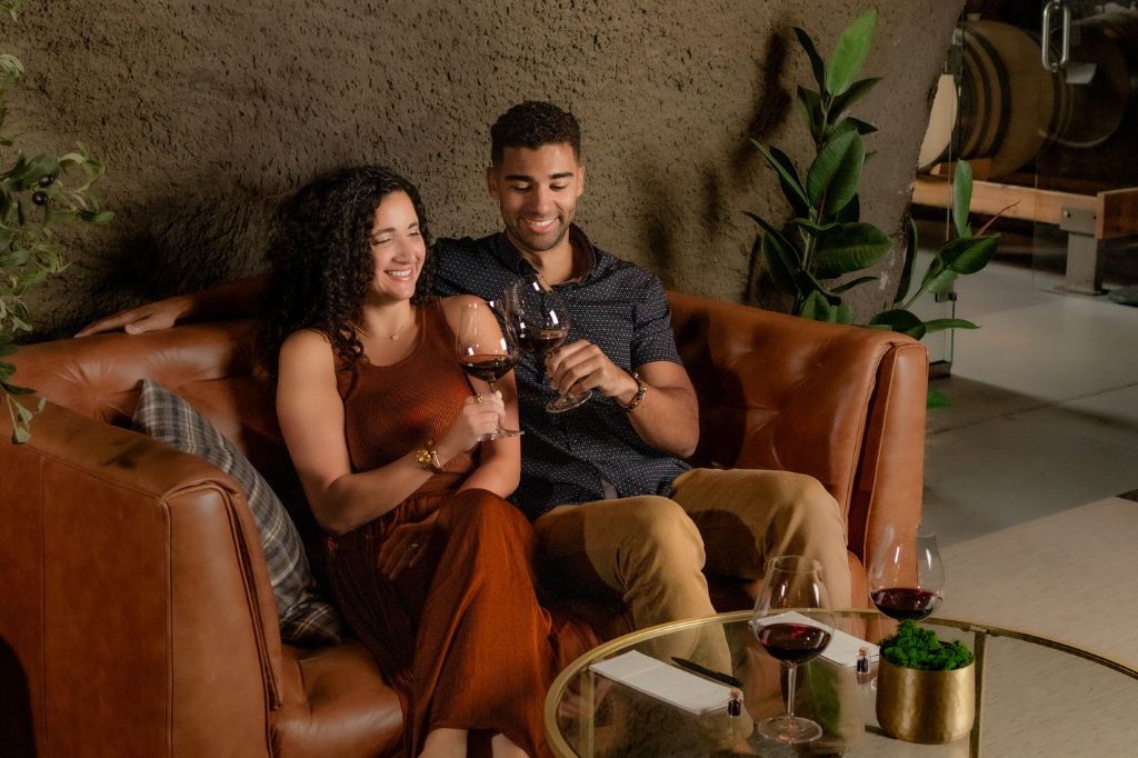A smiling couple sits on a brown leather couch, clinking glasses of red wine. They are relaxed and enjoying each others company, with plants and wine barrels in the background. A table with wine glasses is in front of them.