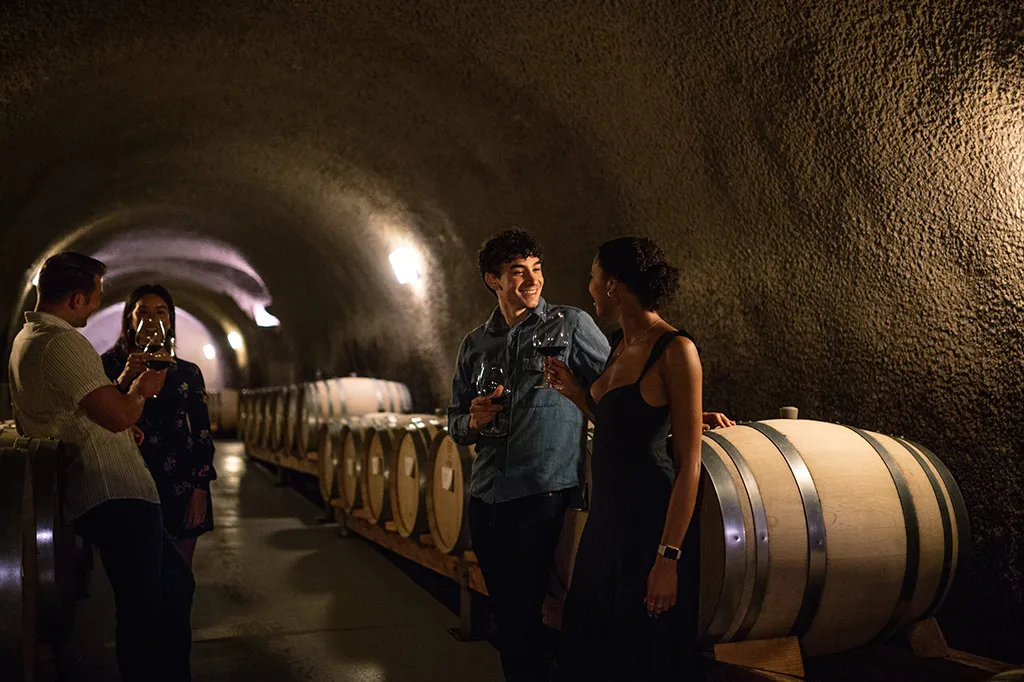 Four people stand in a dimly lit wine cellar, holding wine glasses and talking near wooden barrels lined up along the curved stone wall. Two are facing each other and smiling in the foreground.