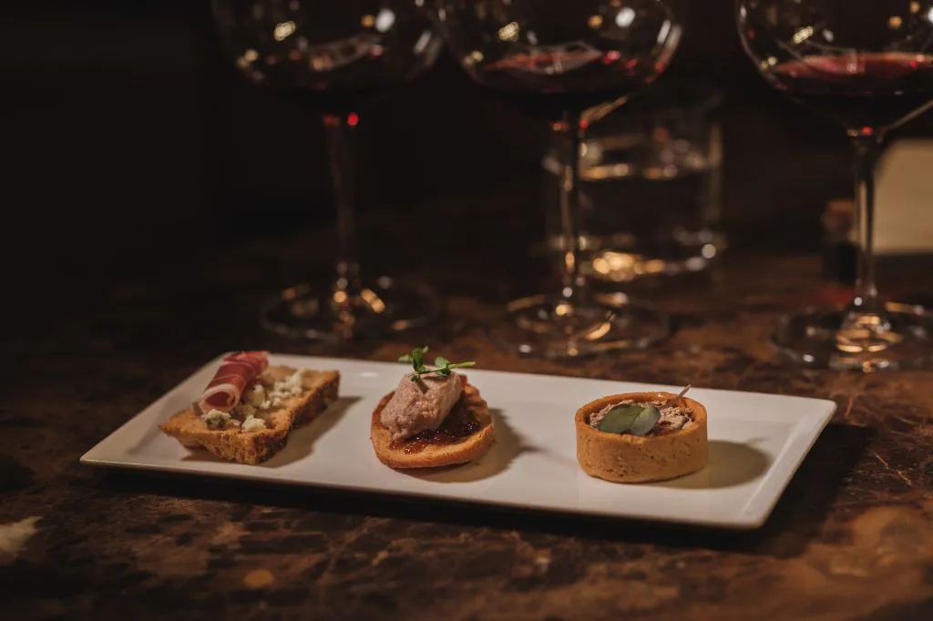 A rectangular white plate with three assorted gourmet appetizers, each on different breads or pastry bases, is set on a dark table beside glasses of red wine.