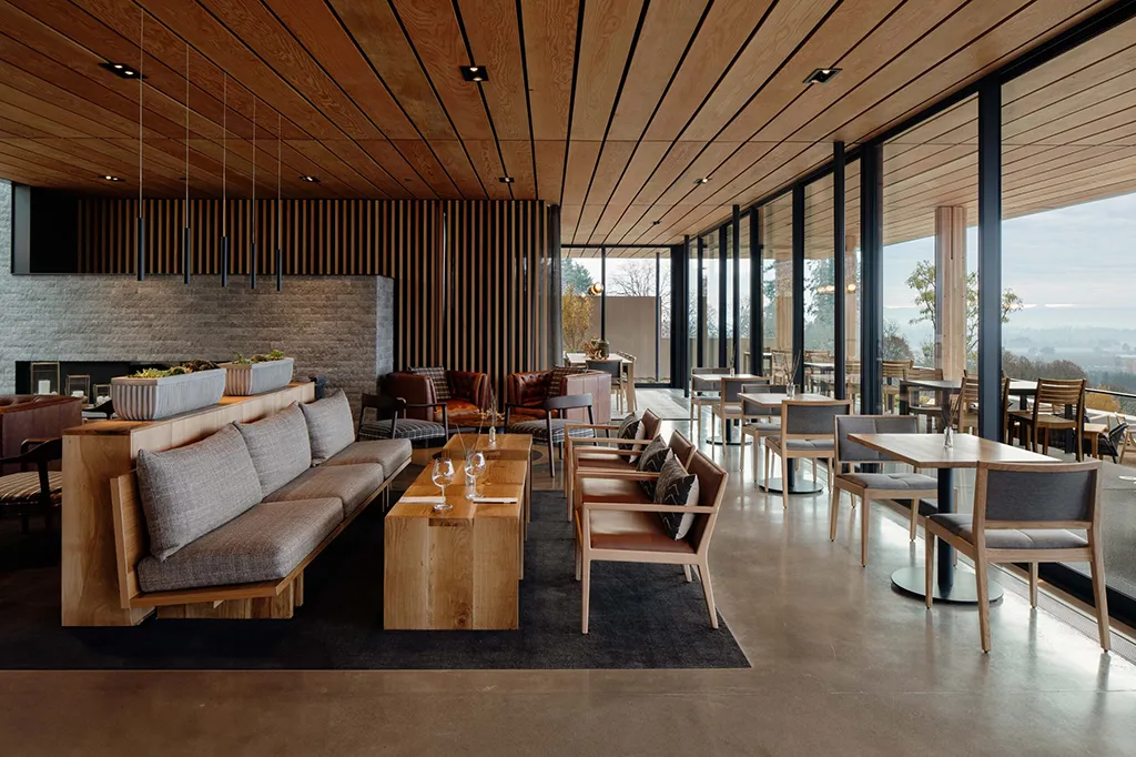 A modern lounge with wood-paneled ceilings, large windows, and minimalist furniture. There are gray cushioned sofas, wooden tables and chairs, and a view of trees and sky outside. The space is spacious and well-lit.