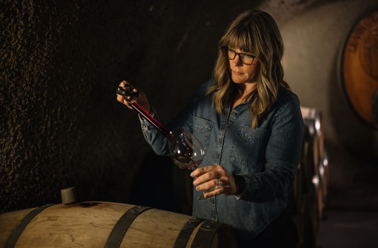 A woman with long hair and glasses, wearing a denim shirt, uses a wine thief to extract red wine from a barrel into a glass in a dimly lit cellar.
