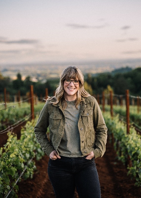 A woman with shoulder-length blonde hair and glasses stands smiling in a vineyard, wearing a green jacket and jeans, with rows of grapevines and a scenic landscape in the background.