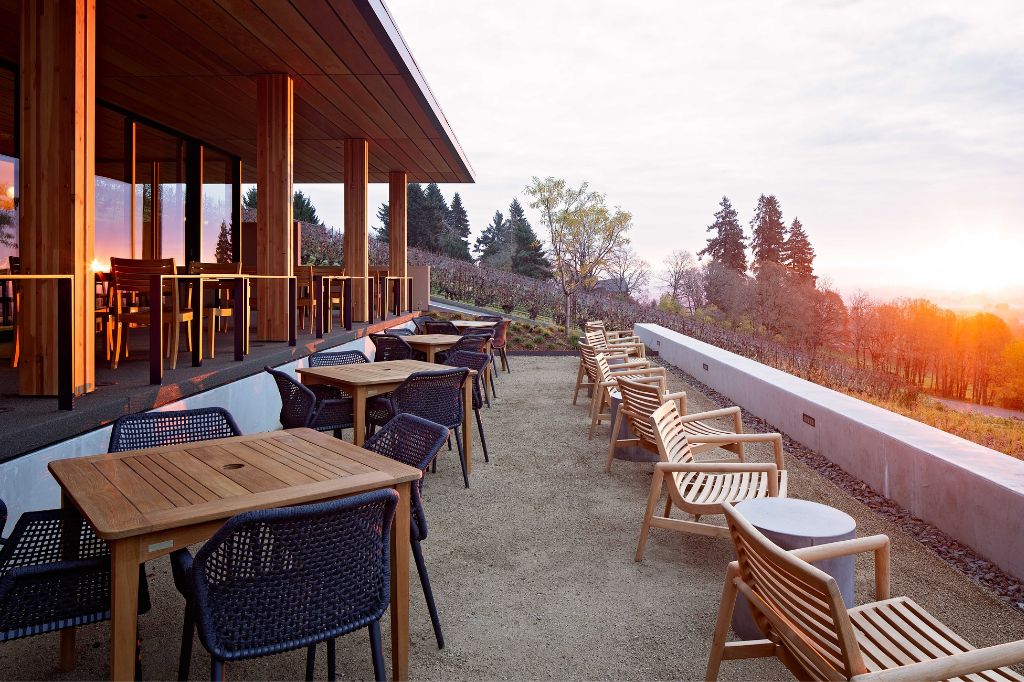 Outdoor restaurant patio with wooden tables and chairs, bordered by a low concrete wall, overlooking a scenic landscape with trees at sunset. The building has large glass windows reflecting the evening light.