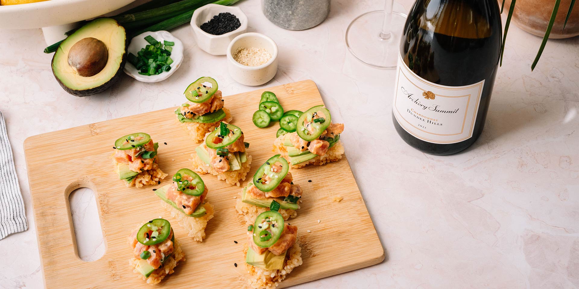 A wooden board with bite-sized sushi stacks topped with avocado, spicy tuna, jalapeño slices, and sesame seeds. A bottle of sparkling wine, two glasses, an avocado, and bowls of sesame seeds and green onions are nearby.