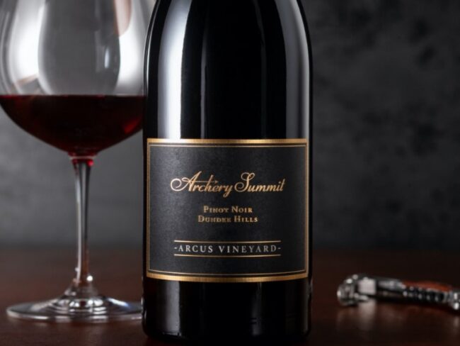 A bottle of Archery Summit Pinot Noir wine labeled Dundee Hills Arcus Vineyard sits next to a filled wine glass and a corkscrew on a dark surface with a blurred dark background.