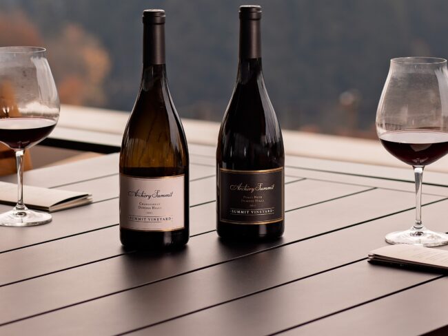 Two wine bottles and two partially filled glasses of red wine are arranged on a dark outdoor table, with scenic, blurred trees and hills in the background.