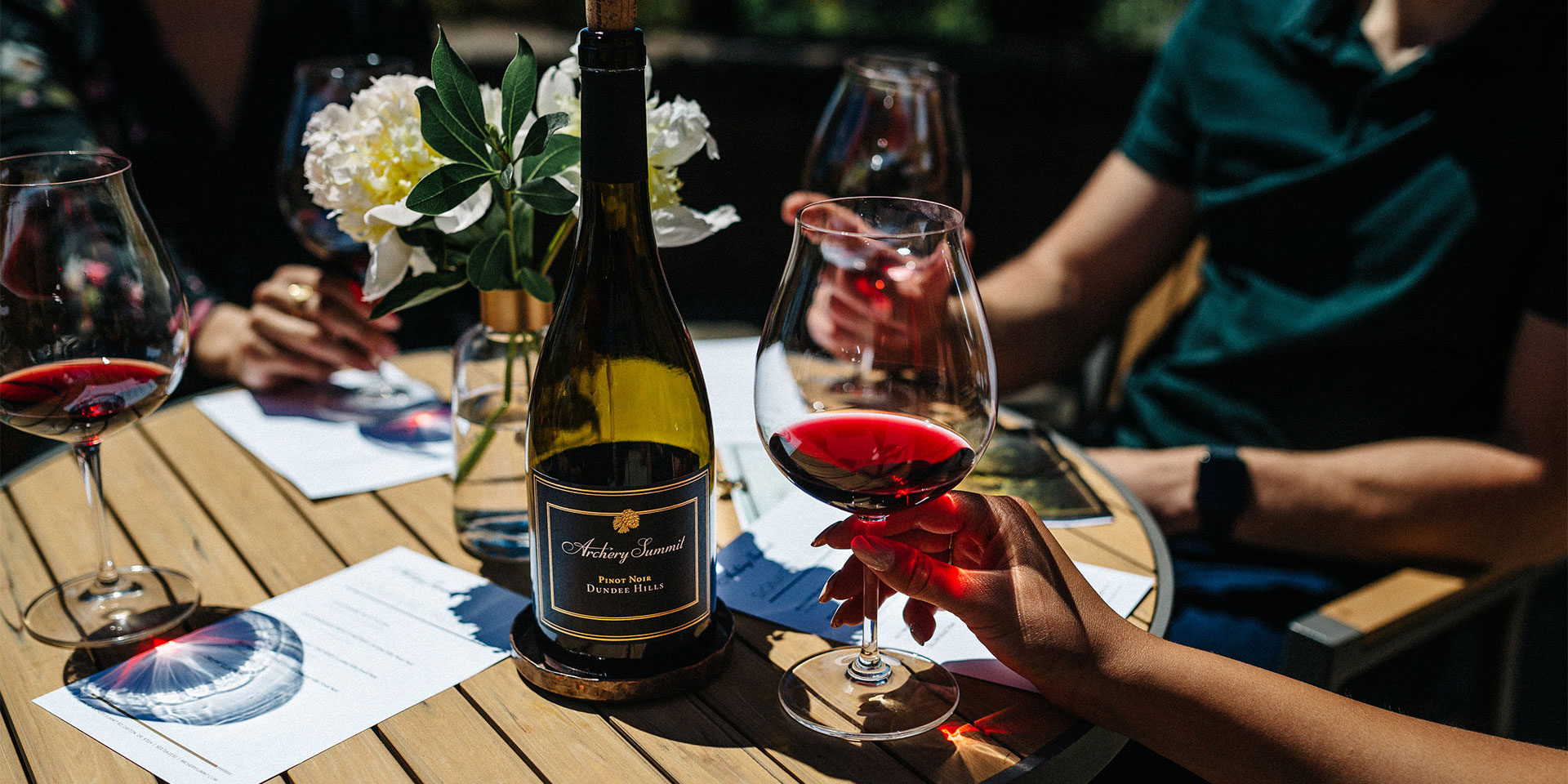 A close-up of a hand holding a glass of red wine next to a wine bottle on a wooden table, surrounded by people, wine glasses, flowers, and tasting notes in an outdoor setting.