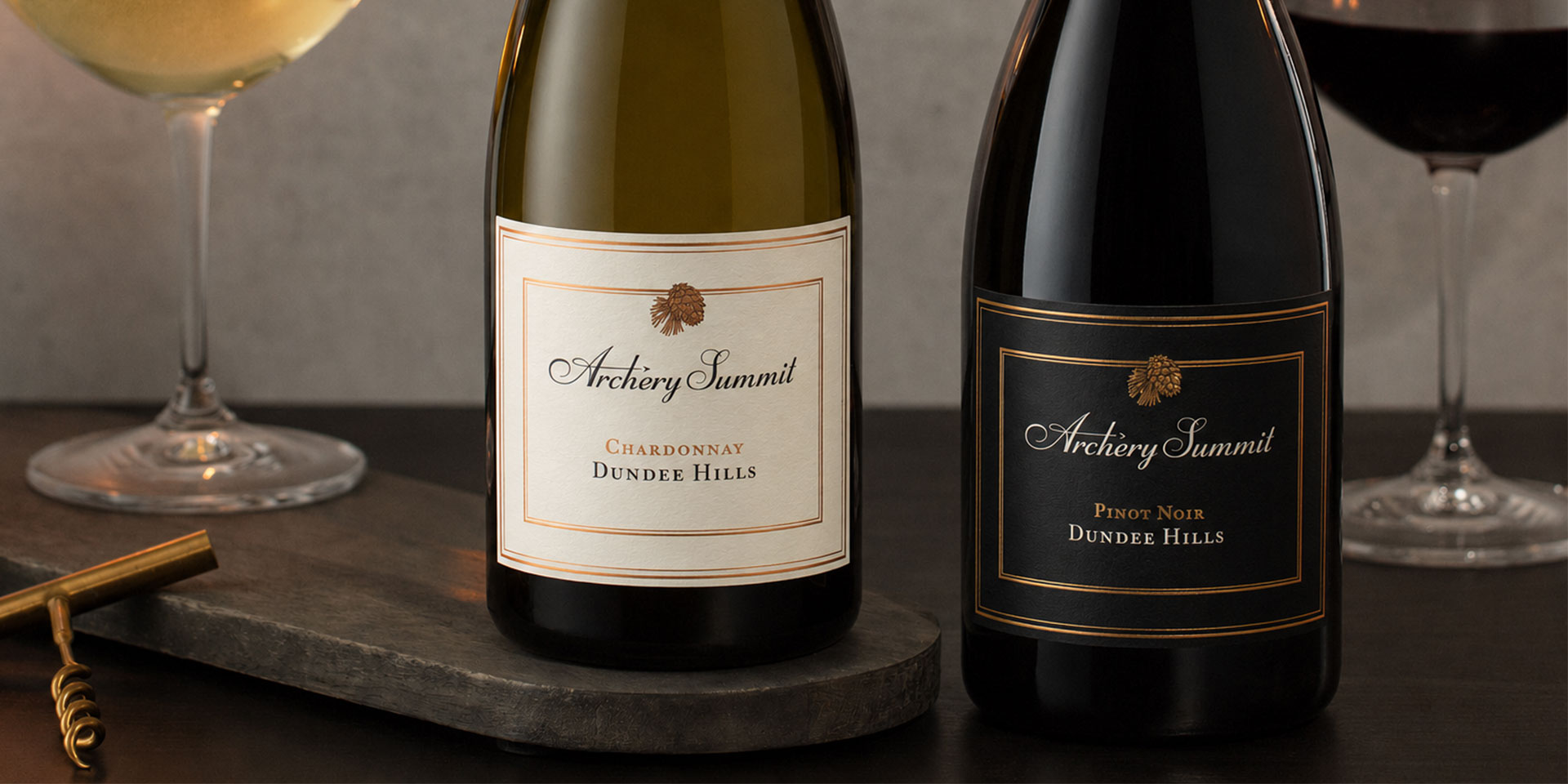 Two bottles of Archery Summit wine, one Chardonnay and one Pinot Noir, are displayed on a dark table with a corkscrew and two wine glasses, one filled with white wine and one with red wine, in the background.