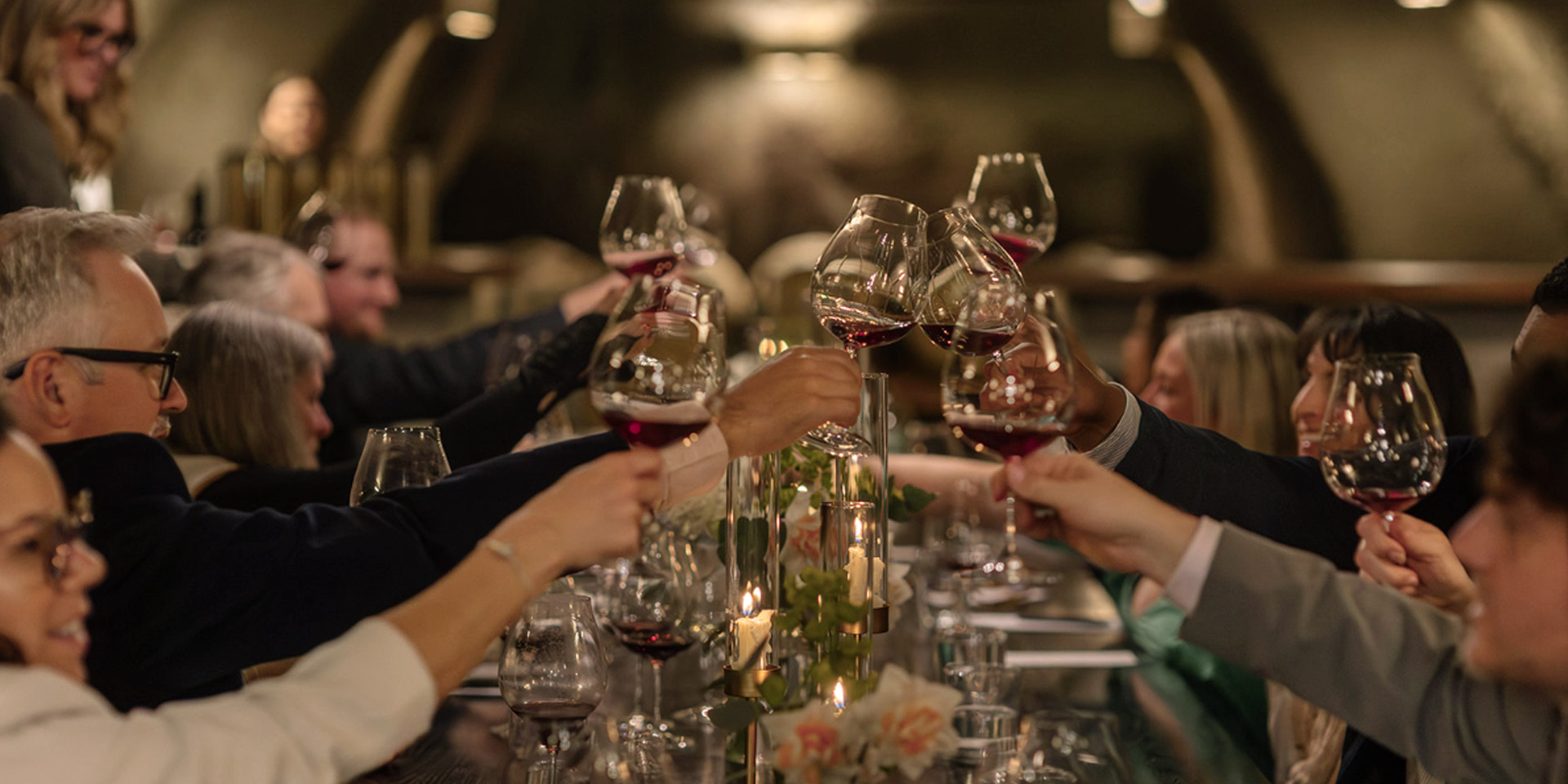 A group of people seated around a candlelit dinner table raise their wine glasses in a toast, creating an atmosphere of celebration in a warmly lit, elegant setting.