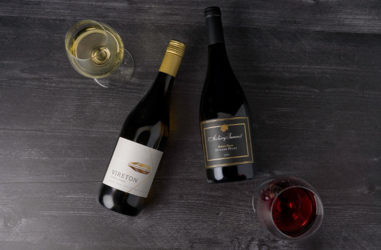 Two bottles of wine, one with a white label and one with a black label, lie on a dark wooden surface next to two glasses—one filled with white wine, the other with red wine.