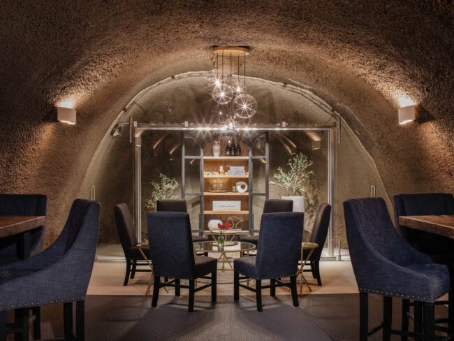 A stylish dining area inside a textured, arched cellar features a round glass table surrounded by six dark upholstered chairs and a modern chandelier overhead, with shelves and decorative plants at the back.