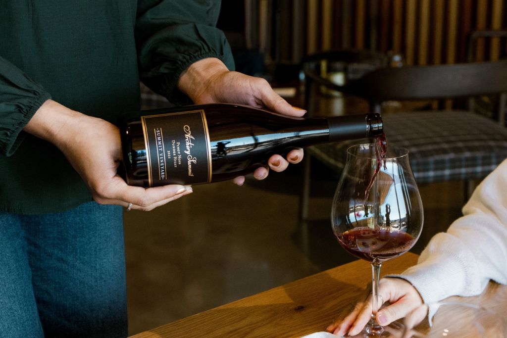 A person pours red wine from a dark bottle into a large wine glass held by another person sitting at a wooden table. The background is softly lit and out of focus.