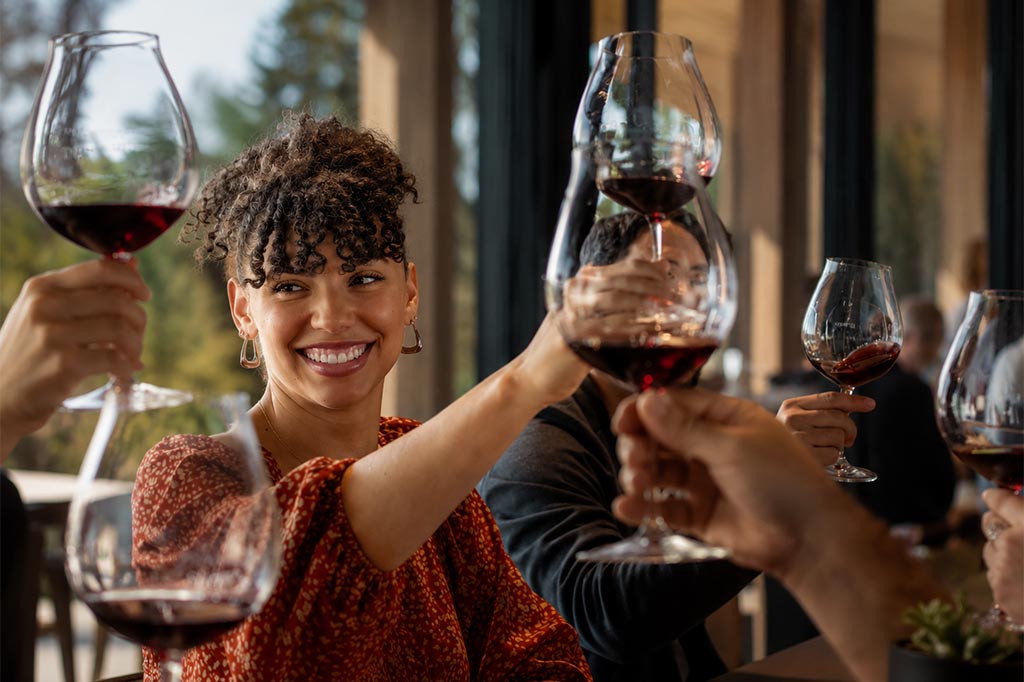 A smiling woman raises a glass of red wine for a toast with friends at a sunlit restaurant, surrounded by people holding large wine glasses. The atmosphere appears joyful and warm.