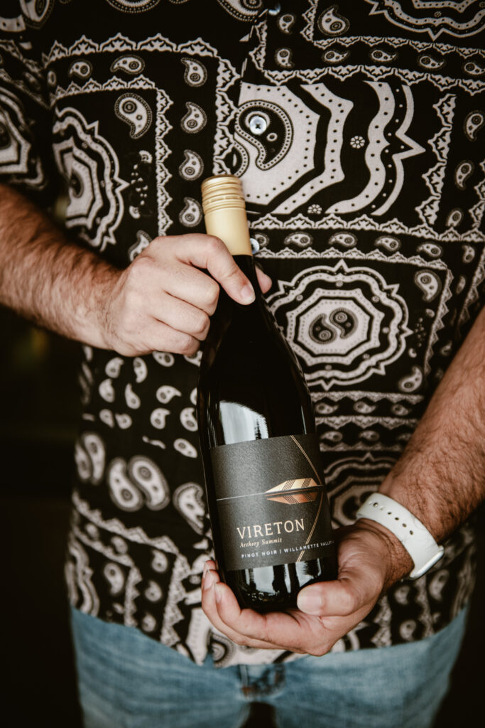 A person in a patterned black and white shirt holds a bottle of wine labeled Vireton with a gold feather design. The person wears a white watch and blue jeans.