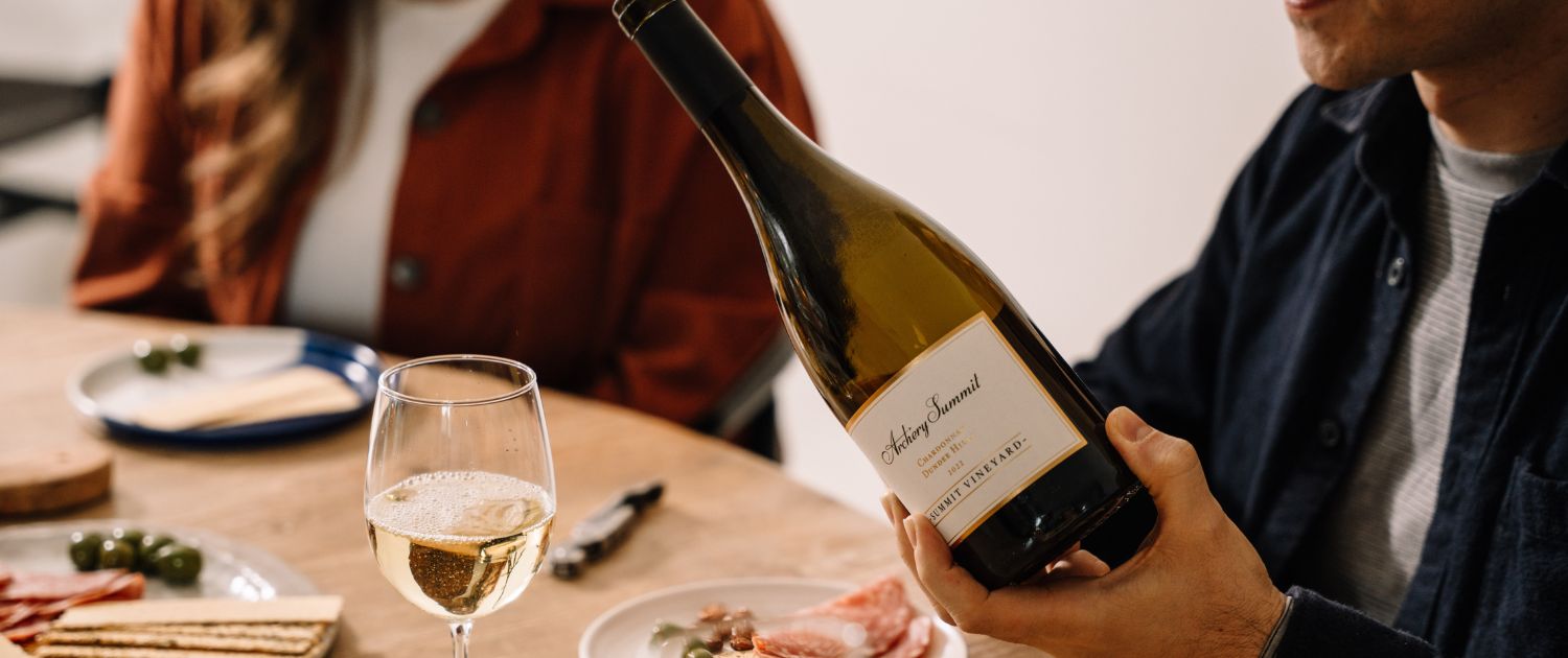 A person holds a wine bottle at a table set with a glass of white wine, plates of cheese, crackers, olives, and charcuterie, while another person sits across from them.